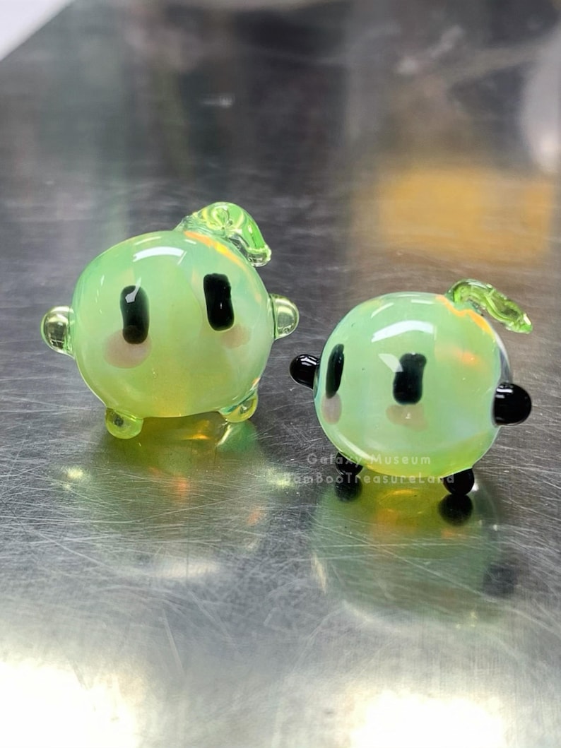 May include: Two small, round, glass figurines in a light green colour. Each has black eyes, pink cheeks, and small black arms and legs. One has a small green leaf on top. The other has a small green leaf on top.