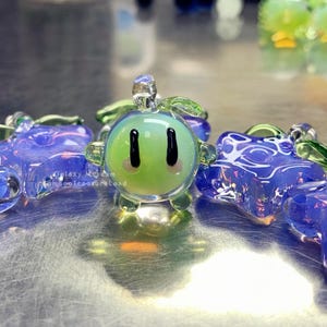 May include: A selection of glass art pieces in various colours. The central piece is a green, spherical figure with black eyes and a smiling face. Other pieces are blue and purple, with detailed patterns. The pieces are displayed on a reflective surface.