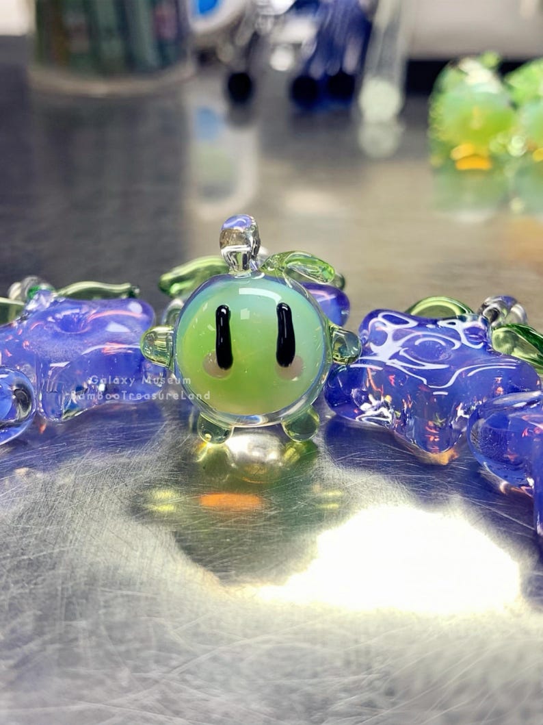 May include: A selection of glass art pieces in various colours. The central piece is a green, spherical figure with black eyes and a smiling face. Other pieces are blue and purple, with detailed patterns. The pieces are displayed on a reflective surface.