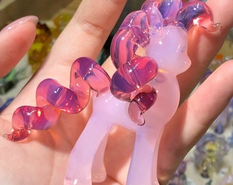 May include: A pink glass figurine of a pony with a flowing mane and tail. The pony has a light pink body with darker pink and purple accents in the mane, tail, and decorative elements. The glass is translucent, allowing light to pass through.