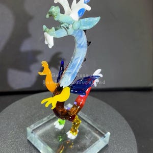 May include: A colorful glass dragon sculpture with a clear square base. The dragon features shades of blue, green, yellow, and red. The sculpture is approximately 6 inches tall and is displayed on a dark gray surface.
