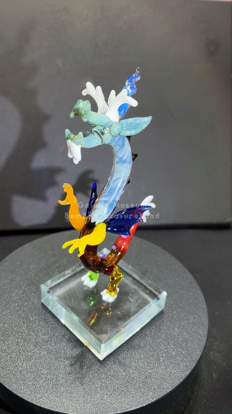 May include: A colorful glass dragon sculpture with a clear square base. The dragon features shades of blue, green, yellow, and red. The sculpture is approximately 6 inches tall and is displayed on a dark gray surface.