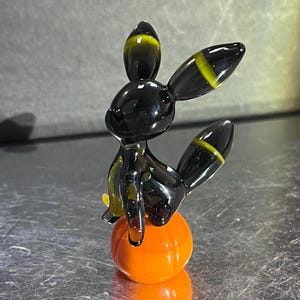 Puede incluir: Una figurita de vidrio negro de un personaje de Pokemon con detalles amarillos, sentada sobre una esfera de vidrio naranja.