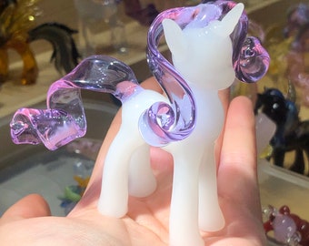 May include: A white glass unicorn figurine with a purple and pink mane and tail. The unicorn is standing on a hand.