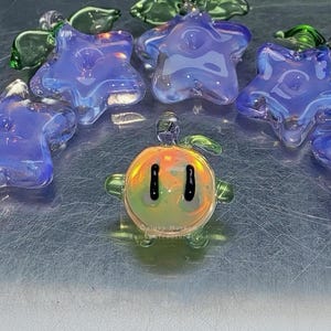 May include: A selection of handmade glass ornaments. The image shows several blue, star-shaped pieces with an iridescent finish, and a small, round, orange and yellow character with black eyes. Green glass angel wings are also visible.