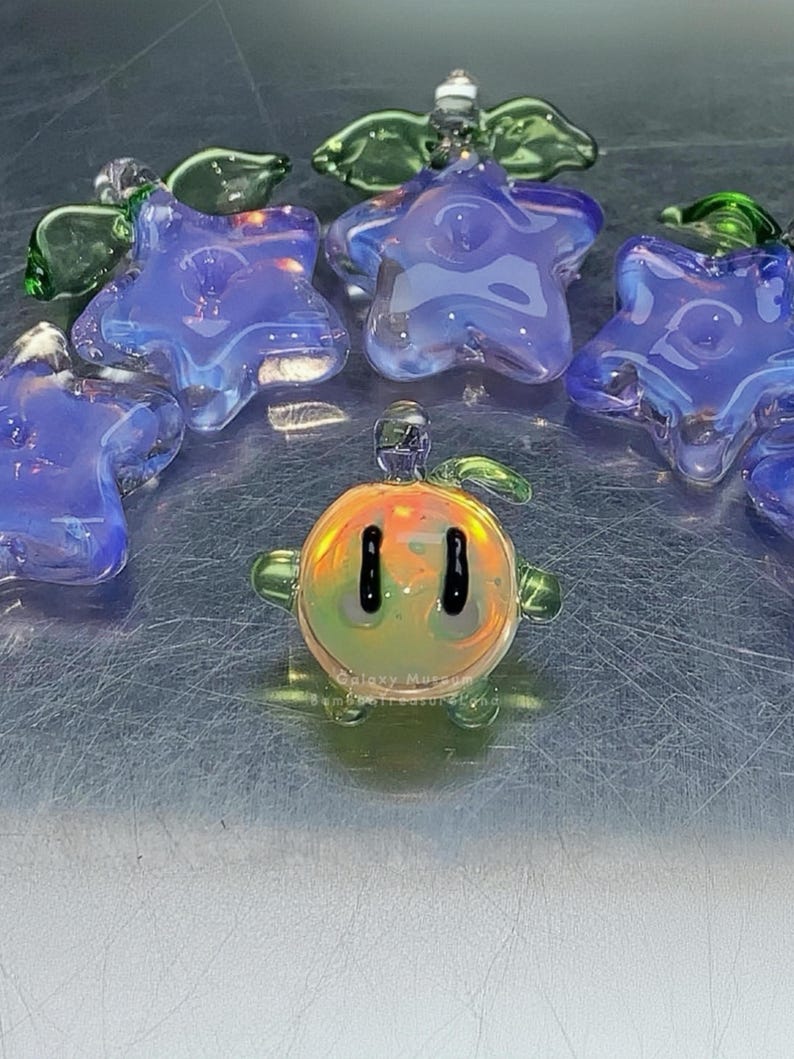 May include: A selection of handmade glass ornaments. The image shows several blue, star-shaped pieces with an iridescent finish, and a small, round, orange and yellow character with black eyes. Green glass angel wings are also visible.