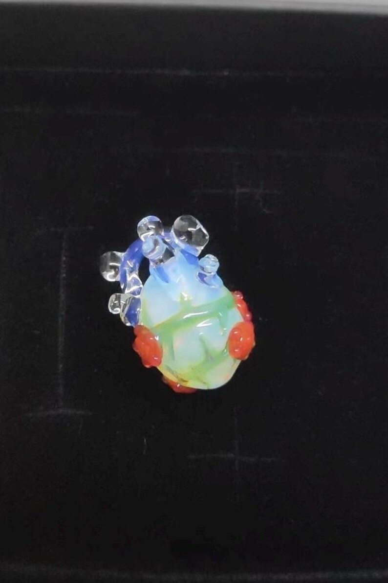 May include: A small glass heart charm with red and blue accents. The heart is translucent and has a green swirl inside.
