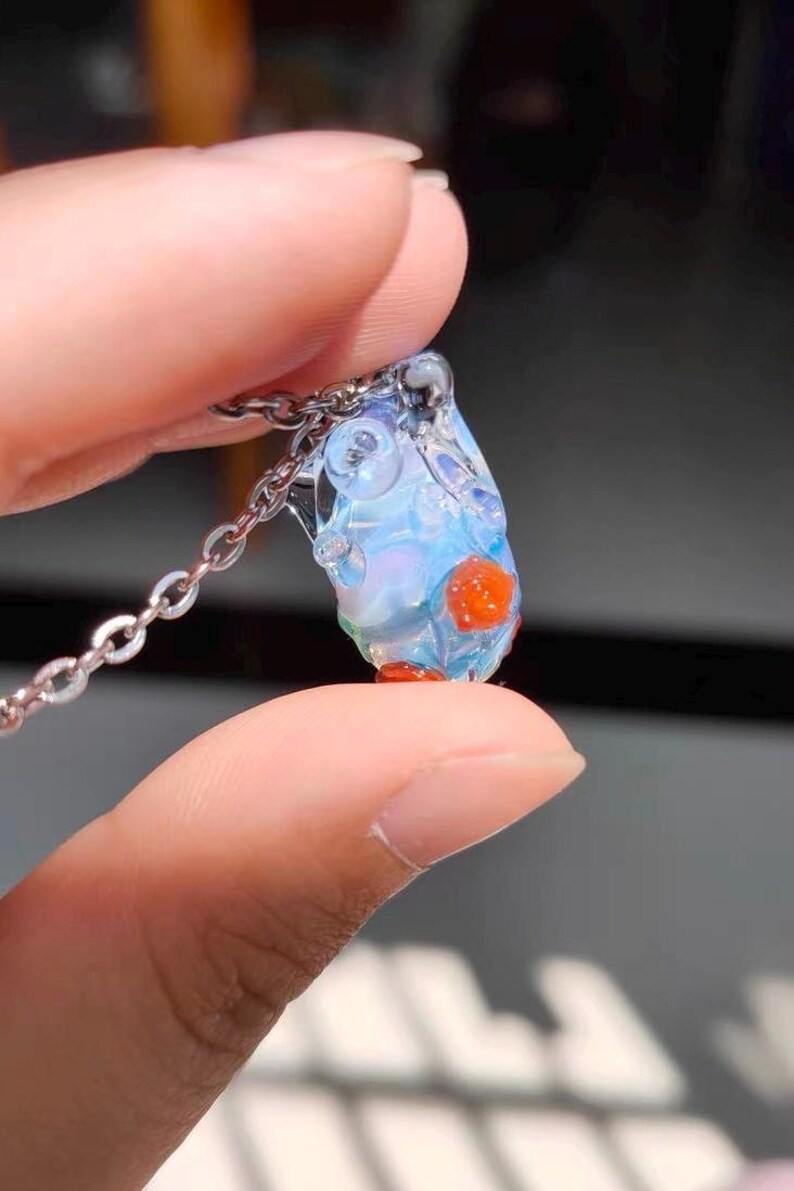 May include: A delicate glass pendant necklace with a clear, iridescent, teardrop-shaped charm. The charm has small, red, glass beads embedded in it.