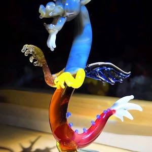 May include: A colorful glass dragon sculpture with a blue, yellow, red, and white design. The dragon has a long, curved body and is mounted on a clear glass base. The dragon's wings and tail are detailed with intricate patterns.