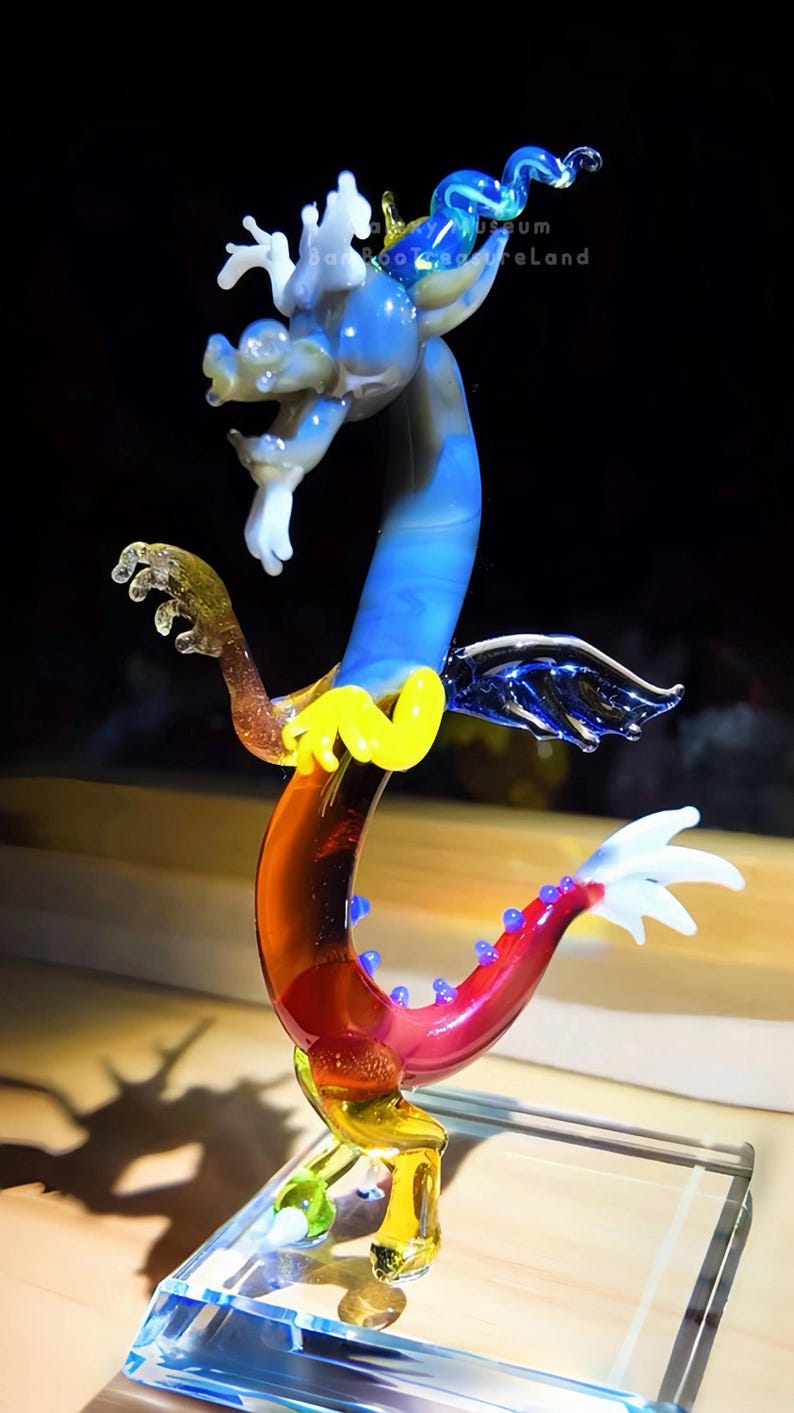 May include: A colorful glass dragon sculpture with a blue, yellow, red, and white design. The dragon has a long, curved body and is mounted on a clear glass base. The dragon's wings and tail are detailed with intricate patterns.