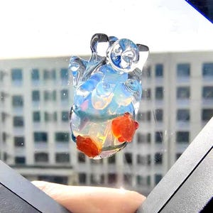 May include: A clear glass heart-shaped ornament with orange and yellow accents. The ornament is suspended in the air, with a blurred background of a building.