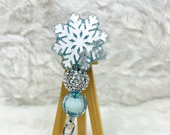 May include: A snowflake-shaped badge reel with a clear, glittery design. The badge reel has a silver rhinestone ball, a light blue bead, and a clear plastic clip. The reel is displayed on a small wooden easel.
