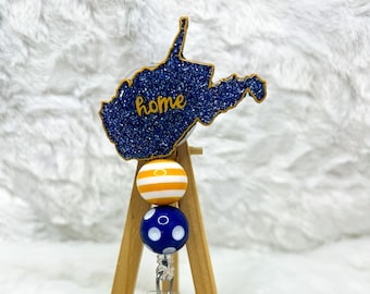 May include: A West Virginia state-shaped badge reel with the word "home" in gold. The badge reel has a blue glitter design and is attached to a wooden easel. The reel features three decorative beads in yellow and blue.