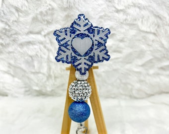 May include: A badge reel with a snowflake design. The snowflake is blue with glitter accents and a white heart in the center. The reel has silver and blue beaded accents and a clear plastic clip.