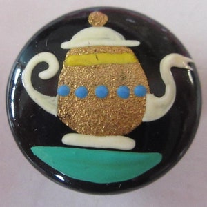 May include: A round, black button with a decorative teapot design. The teapot has a gold glitter body, white spout and handle, and blue dots. The lid and base are white, with a yellow band on the lid and a teal base.