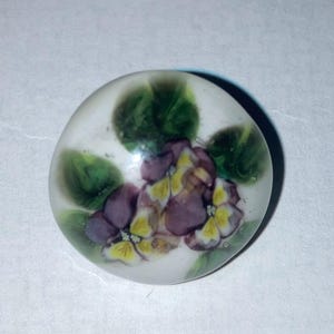 May include: A round, translucent glass button featuring a hand-painted floral design. The motif displays purple and yellow pansy-like flowers surrounded by green leaves, set against a soft, milky white background with a smooth, domed surface.