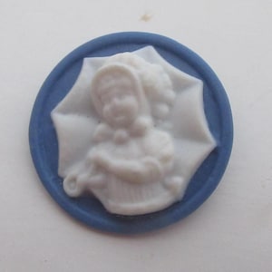 May include: A round, blue and white cameo. The cameo features a white relief of a woman in a bonnet and dress, set against a blue background. The cameo is circular with a raised edge.