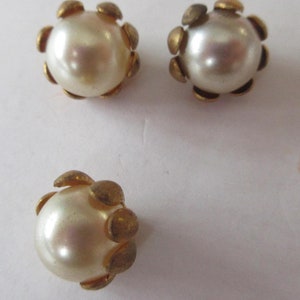 May include: Three vintage pearl-like button earrings. Each earring features a round, off-white pearl-like bead surrounded by six small, gold-colored petal-shaped accents. The earrings are set against a plain, off-white background.