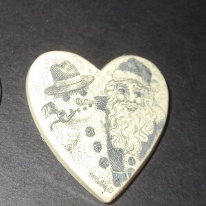 May include: A white heart-shaped ornament with a black and white illustration of a snowman wearing a hat and smoking a pipe, and Santa Claus with a white beard. The ornament is signed "Kynaston".