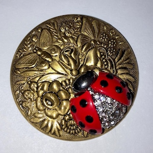 May include: A gold-toned metal button with a raised design of a ladybug, flowers, and butterflies. The ladybug is red with black spots and has clear rhinestones on its wings.