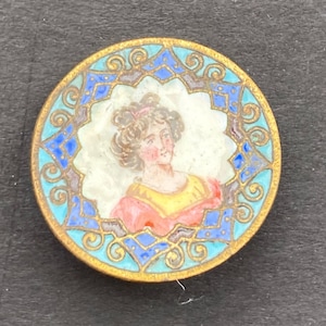 May include: A round, gold-toned button with a blue and white enamel design. The center features a portrait of a woman with brown hair and a yellow and red dress.