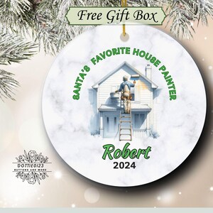 May include: A round ceramic Christmas ornament with a marble effect background. The ornament features a house painter on a ladder painting a house. Text reads "SANTA'S FAVORITE HOUSE PAINTER" and "Robert 2024".