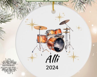 May include: White ceramic ornament featuring a watercolor illustration of a drum set in shades of orange and brown. The name "Alli" and the year "2024" are printed below the drum set. Gold star accents and the text "Free Gift Box" are also visible.