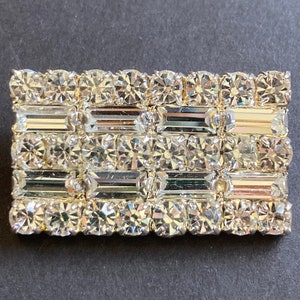 May include: A silver-toned brooch with a rectangular design featuring rows of clear, faceted rhinestones and baguette-shaped crystals.