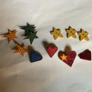 May include: Assortment of wooden buttons in various shapes and colors, including stars, hearts, and a blue heart. The buttons are painted with different patterns and designs.