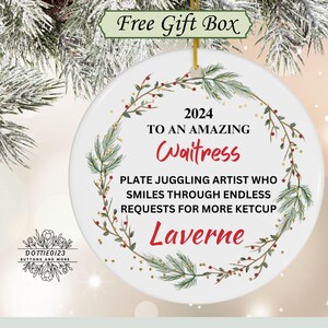 May include: A white ceramic Christmas ornament with a gold string for hanging. The ornament features text that reads "2024 To An Amazing Waitress" and "Laverne". It is decorated with a green and red floral wreath.
