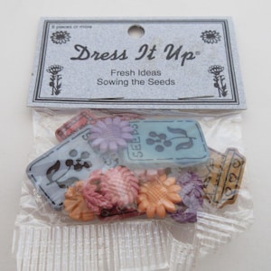 May include: A package of colorful plastic buttons with a floral and gardening theme. The buttons feature images of flowers, seed packets, and gardening tools. The package is labeled "Dress It Up Fresh Ideas Sowing the Seeds".