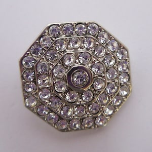 May include: A silver octagonal-shaped brooch with a central clear rhinestone surrounded by concentric circles of smaller clear rhinestones.
