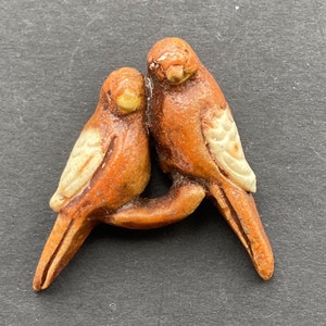 May include: Two brown ceramic parakeets are perched together. The birds have white markings on their wings and tails. The parakeets are facing each other and appear to be cuddling.
