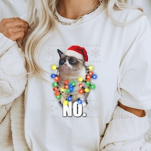 May include: White t-shirt featuring a grumpy cat wearing a Santa hat and Christmas lights. The cat has a displeased expression and the word "NO." is printed below the cat in bold white letters. The shirt has a snowflake pattern.