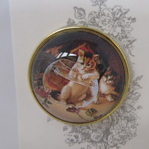 May include: A round, gold-toned pin with a glass dome featuring a vintage illustration of two kittens playing with a glass bowl. The kittens are brown and white with black markings.