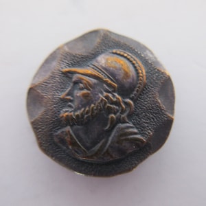 May include: A bronze-colored, circular button with a relief of a man in profile wearing a helmet and beard. The button has a textured, hammered-metal appearance. The man's face and helmet are detailed with fine lines.