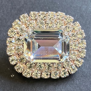 May include: A large, clear, rectangular crystal is surrounded by a border of small, clear rhinestones. The rhinestones are set in a gold-toned metal.