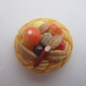 May include: A yellow and orange fruit basket brooch with a woven design. The basket is filled with apples, leaves, and a cinnamon stick.