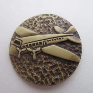 May include: A round, brown, textured button with a beige airplane design. The airplane has three propellers and a row of windows.