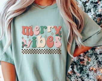 May include: A light green t-shirt with the words "merry vibes" in a retro font. The text is in shades of pink, green, and beige, with holly and snowflake accents. A checkered pattern borders the text.