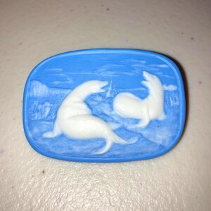 May include: A blue oval cameo featuring two white seals. The seals are in relief against a blue background, with a landscape scene in the background. The cameo is likely made of ceramic or porcelain.