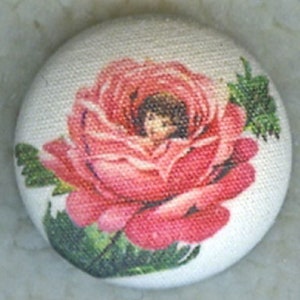 May include: A round, fabric-covered button featuring a detailed illustration of a pink rose with green leaves. The rose has a face in the center. The button has a white background with a textured appearance.