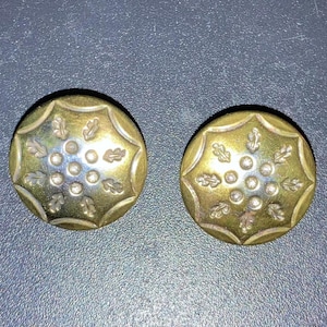 Puede incluir: Dos botones de metal de color dorado con un diseño decorativo. Cada botón presenta un círculo central rodeado por un anillo de círculos más pequeños y un patrón de hojas.