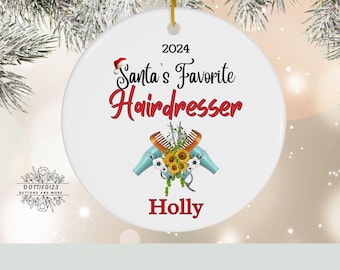 May include: White ceramic Christmas ornament with the text "Santa's Favorite Hairdresser 2024" in black and red. Features a Santa hat, hair dryers, combs, and sunflowers. The name "Holly" is at the bottom.