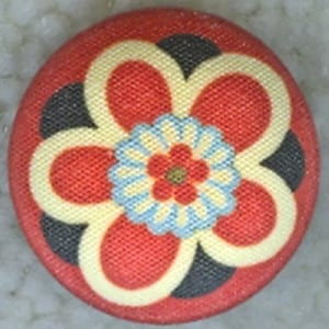 May include: A red, white, and black fabric button with a stylized flower design. The flower has a center with a smaller red flower and blue and white petals.