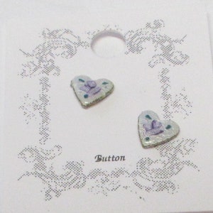 May include: Two heart-shaped buttons with a floral design in shades of purple, green, and white. The buttons are on a white card with the word "Button" printed on it.