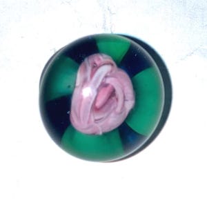 May include: A spherical glass paperweight featuring a clear outer layer with alternating dark blue and bright green vertical segments. Encased within the center is a pale pink, swirling floral-like design with soft, layered petal textures.