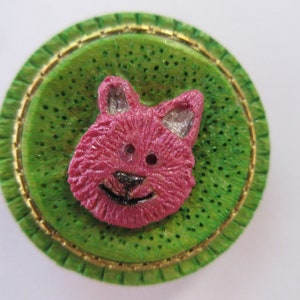 May include: A pink cat face button with a green background and a gold trim. The button is round and has a textured surface.