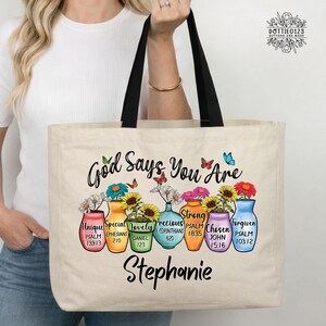 May include: Beige tote bag with black handles featuring the text "God Says You Are" and colorful vases with flowers. Each vase has a word like "Unique" and a Bible verse reference. The name "Stephanie" is printed at the bottom.