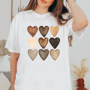 以下が含まれることがあります： 白のTシャツ。水彩画の茶色、ベージュ、タンの色合いの9つのハートのグリッドが特徴です。ハートは3列3段に配置されています。Tシャツは半袖で、ゆったりとしたフィット感です。
